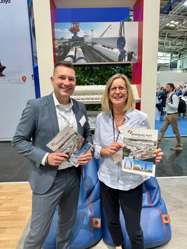 Ein Mann und eine Frau auf einem Messestand. Sie halten Werbebroschüren in den Händen und lächeln in die Kamera.