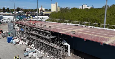 Luftaufnahme des rund 118 Meter langen Stahl-Brückenelements auf der Hafenfläche, umgeben von Gerüstkonstruktionen, Baumaterialien und Arbeitskräften; im Hintergrund sind Industriegebäude und der Nord-Ostsee-Kanal zu sehen