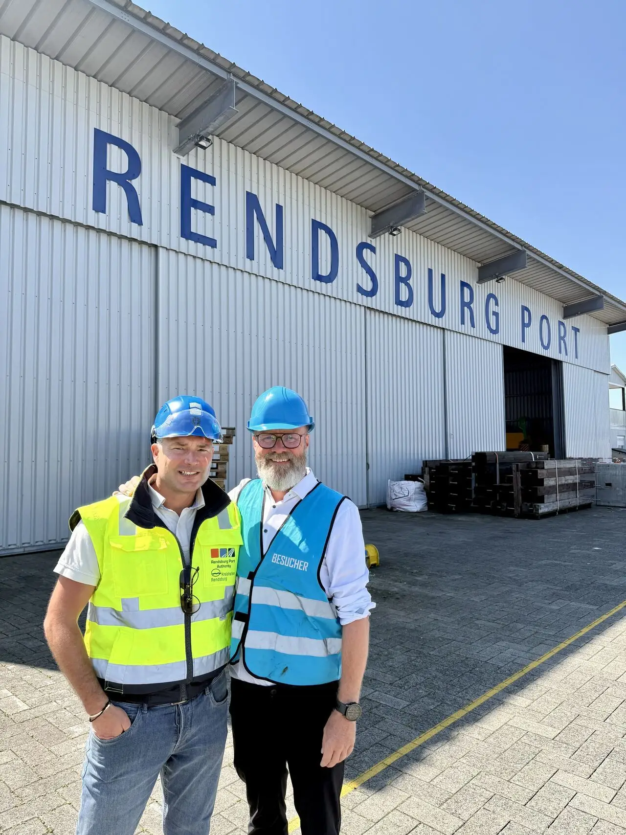 Wirtschaftminister Madsen und WFG-Geschäftsführer Lass stehen in Warnwesten und mit Schutzhelm vor einer großen Halle mit der Aufschrift "Rendsburg Port"