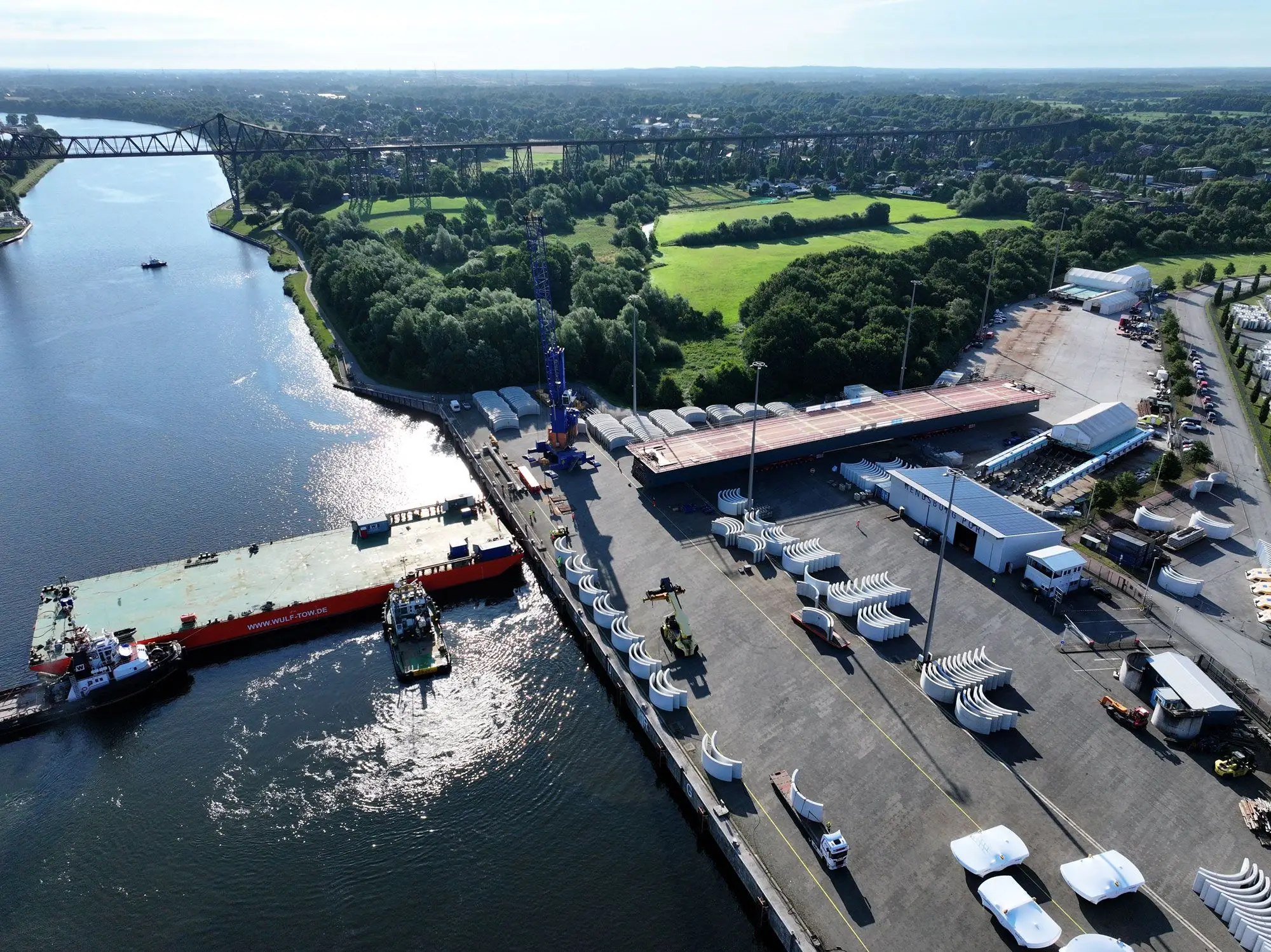 Luftaufnahme: Der Ponton ist quer im Nord-Ostsee-Kanal positioniert. Parallel dazu steht das Mittelstück der Brücke auf der Hafenfläche und wird mittels Schwerlastmodule auf den Ponton verladen