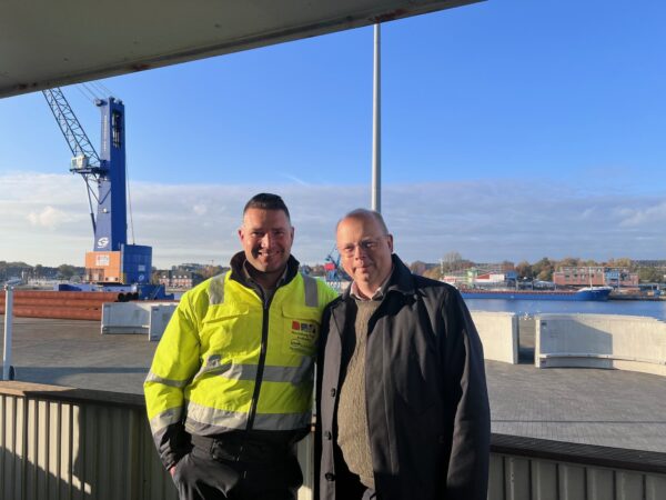 Zwei Manner stehen nebeneinander und schauen in die Kamera. Der linke Mann trägt eine neongelbe Arbeiterjacke, der rechte Mann eine Brille und einen schwarzen Mantel. Im Hintergrund ist Wasser und ein Hafen samt Kran zu sehen.