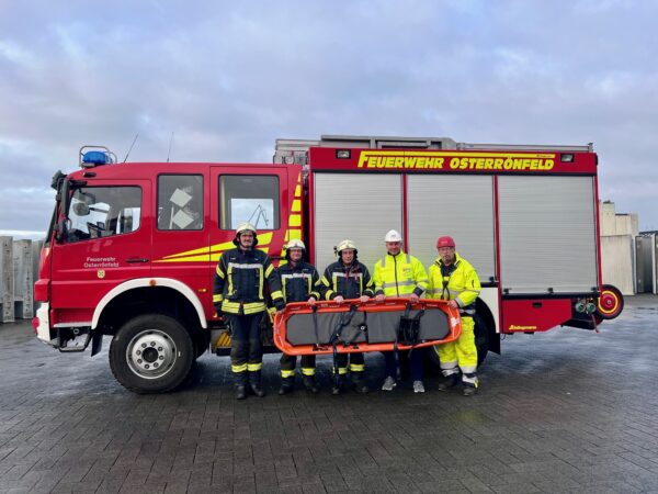 Auf dem Bild ist ein Feuerwehrwagen der Freiwilligen Feuerwehr Osterrönfeld zu sehen. Davor stehen drei Feuerwehrmänner in voller Montur. Daneben zwei Männer in neongelber Arbeitskleidung mit Helm auf. Alle halten eine Schleifkorbtrage, die zur Personenrettung genutzt werden.